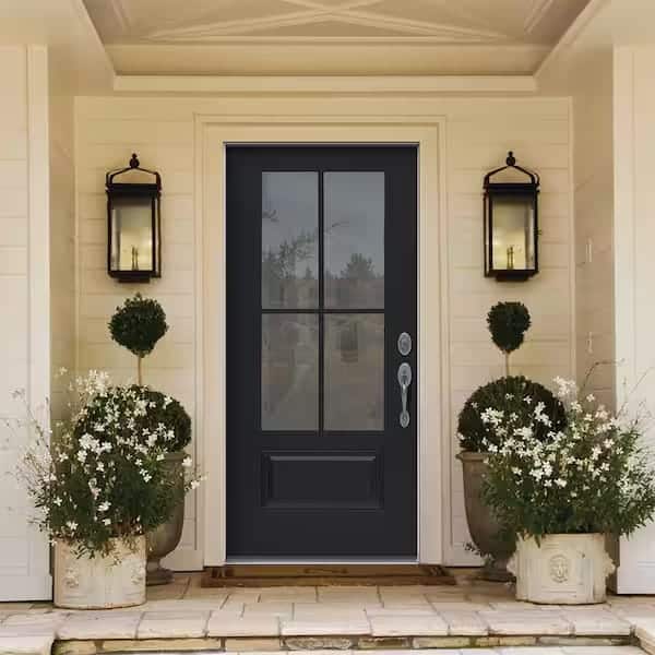 A black front door with glass panels is flanked by two topiary plants in decorative pots and white flowers. Two black lantern-style wall lights hang on either side against a light-colored house exterior.